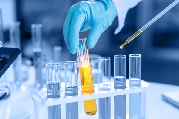 Una mano guantata versa un liquido giallo da una provetta in uno scaffale di provette trasparenti, mostrando le tecniche di laboratorio per l'analisi chimica.