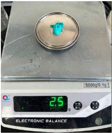 Eine elektronische Waage zeigt eine Probe mit einem Gewicht von 2,5 g. Die Probe ist eine leuchtend türkisfarbene Masse.