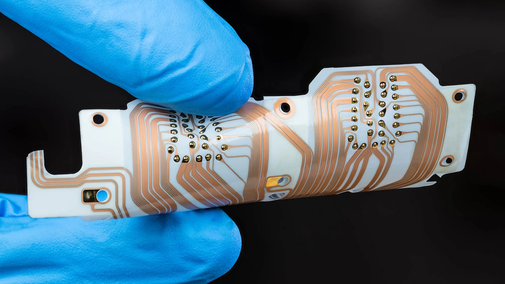 A person with blue gloves holds a circuit board showcasing intricate copper pathways and soldered connections, emphasizing precision in technology.