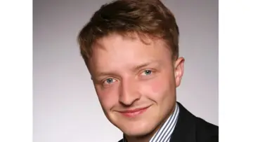 Szemüveges, kék kockás inget viselő üzletember, aki semleges háttér előtt magabiztosan a kamerába néz.