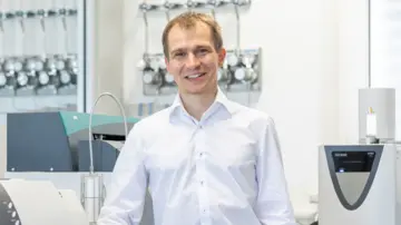 Profesionální muž s brýlemi v modré kostkované košili, který se sebevědomě dívá do kamery na neutrálním pozadí.