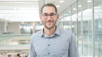 Profesionální muž s brýlemi v modré kostkované košili, který se sebevědomě dívá do kamery na neutrálním pozadí.