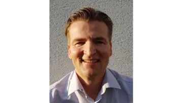 Een zakelijke man met een bril, gekleed in een blauw geruit overhemd, die zelfverzekerd in de camera kijkt tegen een neutrale achtergrond.